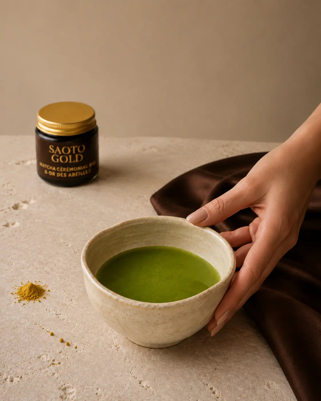 Bol artisanal de matcha cérémonial Saoto Gold tenu à la main, avec le pot Saoto Gold flouté en arrière-plan sur une pierre ivoire et un tissu brun, pollen de fleurs à côté du bol